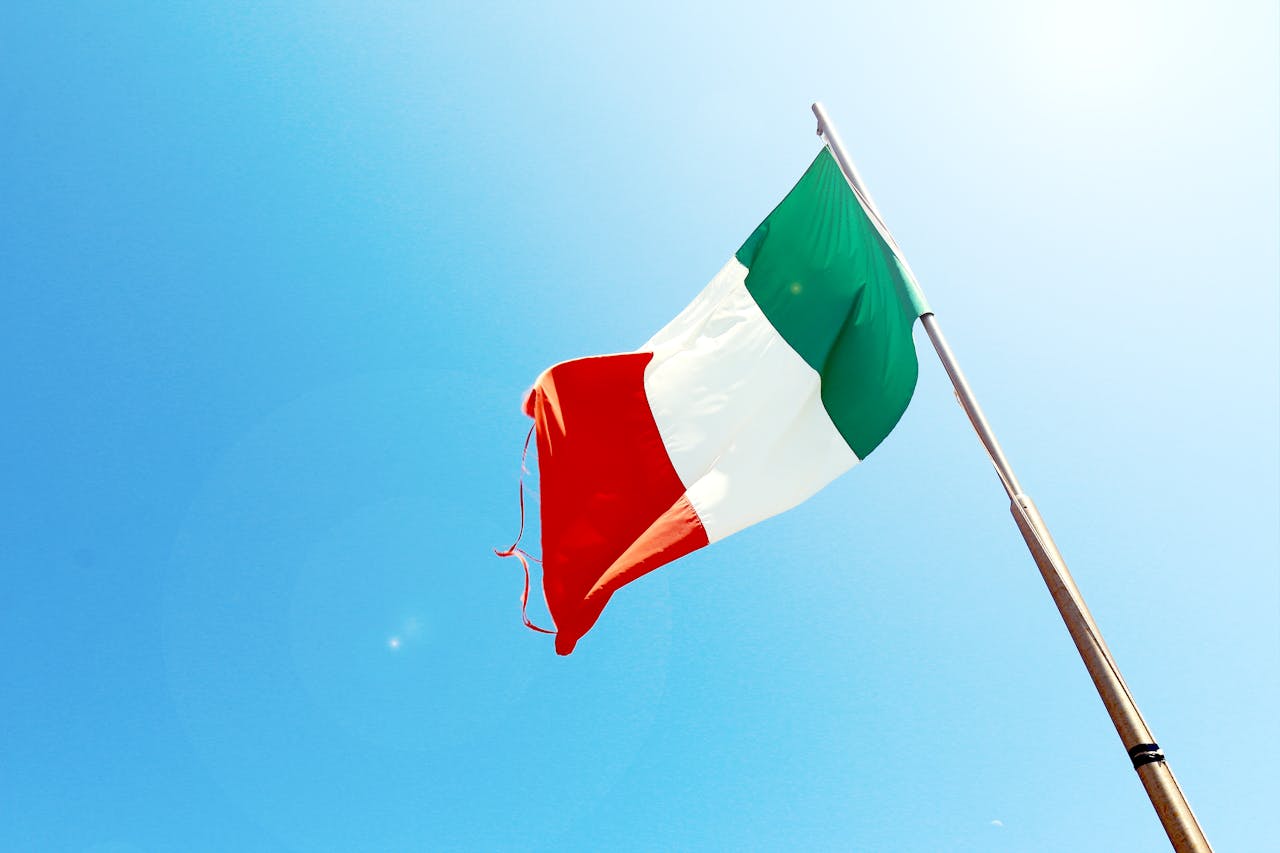Afbeelding van een vlaggenstok met italiaanse vlag met een blauwe lucht als achtergrond en een felle zon.