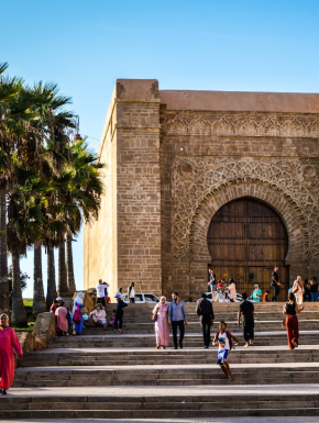 Afbeelding van een bruine betonnen trap waarop mensen lopen richting een bruin betonnen gebouw.